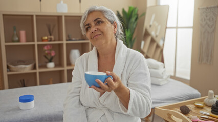 Mature grey-haired woman in a white robe holding a blue cup in a spa room with shelves and wellness...
