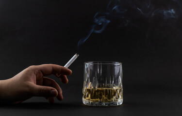 A burning cigarette in one hand and a glass of alcohol on the black background.