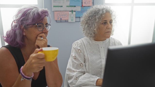Mature women working together in an office, one holding a coffee cup, seriously focused on the computer screen.