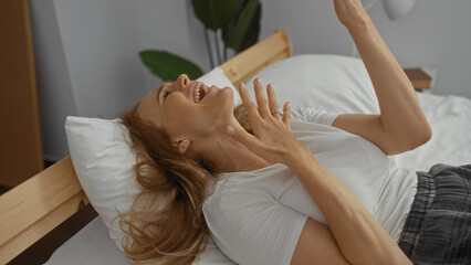 Woman laughing on bed in relaxed home setting with blonde hair wearing casual wear indoors in a sunlit bedroom creating a serene happy atmosphere.