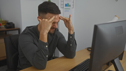 Hispanic man sitting indoors at an office desk, looking stressed while touching his temples in...