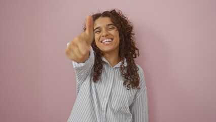 Beautiful young woman with brunette hair smiling and showing thumbs up, isolated over a pink...