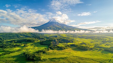Fototapeta premium Majestic volcano rises above lush green fields under a clear blue sky, surrounded by wispy clouds, showcasing nature's breathtaking beauty.