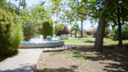 Blurred image of a sunlit outdoor park with a defocused fountain surrounded by greenery and trees on a bright day