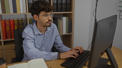 Hispanic man working at a computer in an office setting with a beard and glasses, surrounded by...