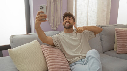 Young man indoors sitting on a couch giving a thumbs up gesture while taking a selfie in a comfortable living room setting