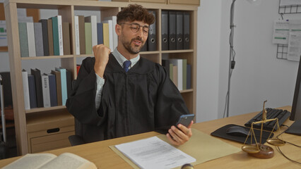 Young, handsome, man in a judge's robe celebrating in an office, using a smartphone, with legal bookshelves and scale in the background.