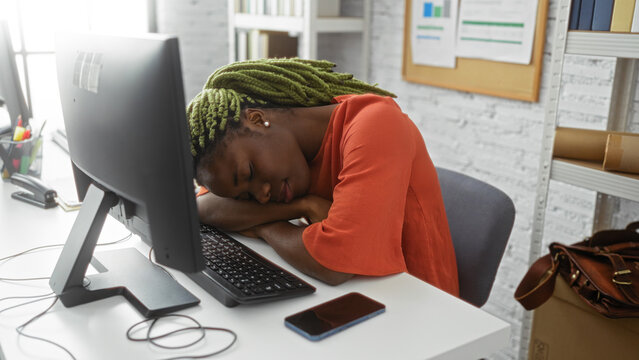 Woman resting at desk inside office with head on arms wearing orange shirt beside computer, phone on desk emphasizes workplace fatigue and relaxation, young adult setting. - Powered by Adobe