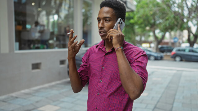 Young black man talking on phone while holding cigarette on urban street background outdoors.