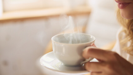 Close-up shot of woman with steaming cup of coffee, capturing warmth and relaxation in softly lit room. Concept of wake up, beauty, weekend, morning routine, home.
