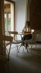 Young woman with glasses sitting at a small table in a cozy indoor cafeteria, looking out the window with natural light illuminating the room.