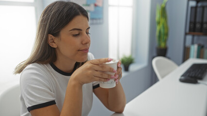 Young woman enjoying coffee break in office room while sitting at desk holding a cup in her hands