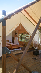 A young hispanic brunette woman enjoys a beautiful outdoor camping experience in africa, standing inside a tent with elegant furnishings and wooden elements under a clear blue sky.