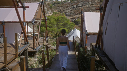A young hispanic brunette woman explores a beautiful camping site with wooden tents outdoors in africa, strolling along the pathways under the warm sun, immersed in nature.
