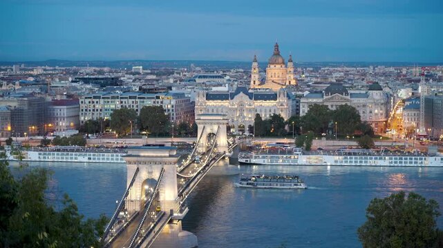 Budapest, Hungary - The famous Szechenyi Chain Bridge (Lanchid) and St. Stephen's Basilica at dusk, with street lights glowing and boats gently drifting on the Danube River