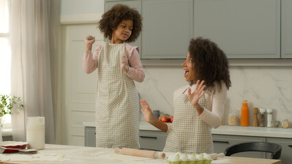 Happy African American family woman female mother mom together fooling fun at kitchen little child kid girl daughter dancing listening music dance in aprons cooking pause relax joy moving weekend