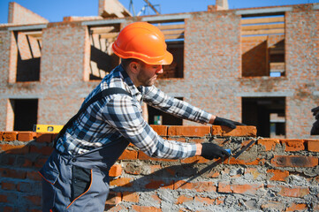 Male construction worker in uniform and helmet on construction of red brick house
