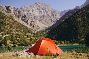 A tourist tent stands by a mountain lake against a background of rocks and peaks. Camping in the mountains in summer © Akhtiamova
