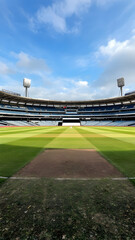 Empty cricket stadium under a bright blue sky. The pitch is visible, with stands largely unoccupied. A tranquil scene.