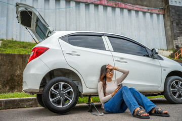 A woman sitting next to her car with a flat tire, looking stressed and using her phone.