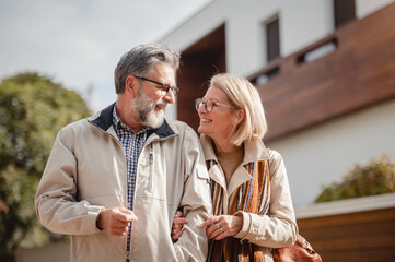 Senior couple embracing and smiling in front of a modern house on a sunny day, showcasing warmth,...