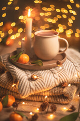 Steaming cup of hot beverage sits on a tray with a mandarin orange and lit candles, placed on a cozy knitted blanket with warm, inviting lighting, creating a perfect winter atmosphere