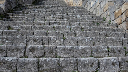An ancient staircase made of large gray stone blocks. The background is made of stone blocks. The steps of the ancient staircase leading to the fortress. Grass grows between the stone blocks.