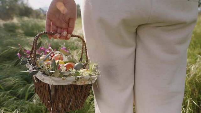 A Woman in White Pants Walks Through a Lush Green Field, Holding a Woven Basket Filled with Colorful Flowers and Easter Eggs. Slow motion.