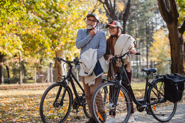 Fototapeta premium Senior couple standing next to their bikes in an autumn park, wearing protective helmets and smiling warmly while enjoying the scenic outdoors