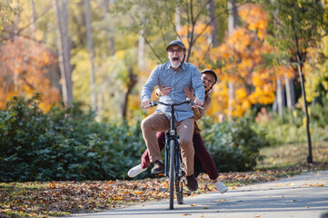 Smiling senior couple riding a bike in a sunny autumn park, wearing protective helmets. The man pedals while the woman enjoys the ride sitting on the rear rack