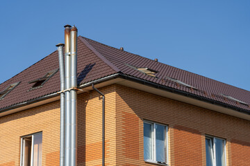 A two-story residential building features a brick facade, sloped roof, and silver chimneys, set against a bright blue sky on a sunny day