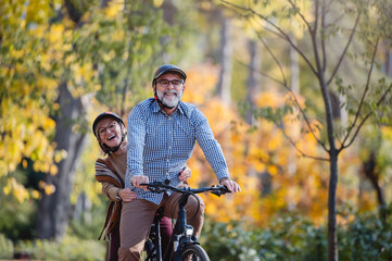 Smiling senior couple riding a bike in a sunny autumn park, wearing protective helmets. The man pedals while the woman enjoys the ride sitting on the rear rack