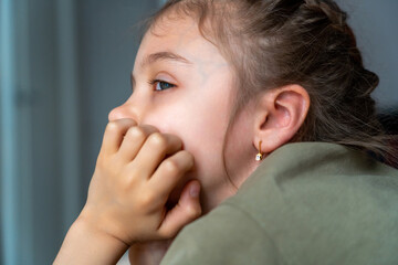 A girl sits quietly with her hand on her face, lost in thought while looking outside, caught in a moment of reflection at home