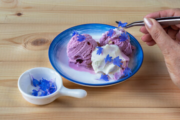 Close-up of hand with tweezers decorating ice cream with raw cornflower petals. Bachelors button. Centaurea cyanus. Ice cream of two colors on saucer with blue border, sprinkled with cornflower petals