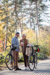 Fototapeta premium Senior couple standing next to their bicycles in an autumn park, looking at a phone together, sharing a moment of connection and curiosity during an outdoor adventure