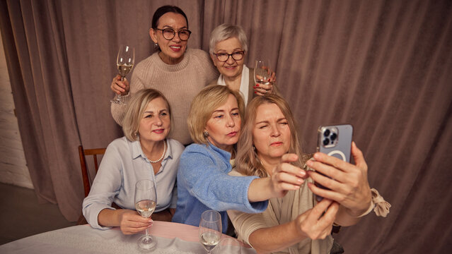 Group of five elderly women, friends, colleagues sitting at home, drinking wine and taking selfie with smartphone. Memorable cozy moment. Concept of elderly lifestyle, positive emotions, celebration