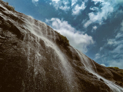 Splashing waterfall on rock mountain with clouds sky in background, low angle view of splashing droplets of stram fresh water from the hill