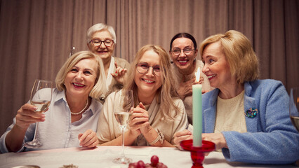 Group of five elderly smiling woman, gathering around table decorated with candles in cozy evening, laughing, talking and drinking wine. Concept of elderly lifestyle, positive emotions