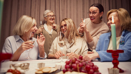 Happy Mothers Day. Elderly women, friends gathering at home, in cozy, warmly-lit living room, laughing, talking and drinking wine. Concept of elderly lifestyle, positive emotions