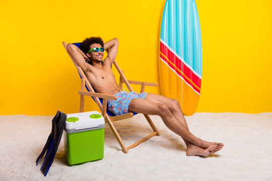 Young man relaxing on beach chair by a colorful surfboard with a yellow background, wearing sunglasses and swim shorts.