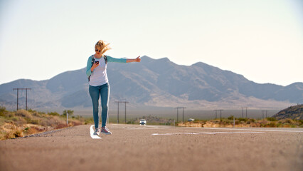 Young Woman Hitchhiking Along Country Road Through Rocky Landscape Looking At Mobile Phone 