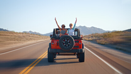 Friends On Road Trip Driving Convertible Car On Country Road With Women Sitting In Back Waving