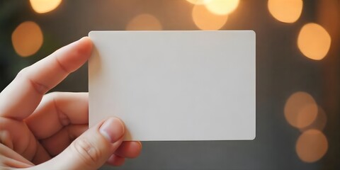 Obraz premium A hand holding a blank white card against a blurred background with warm, glowing lights. Space for text.