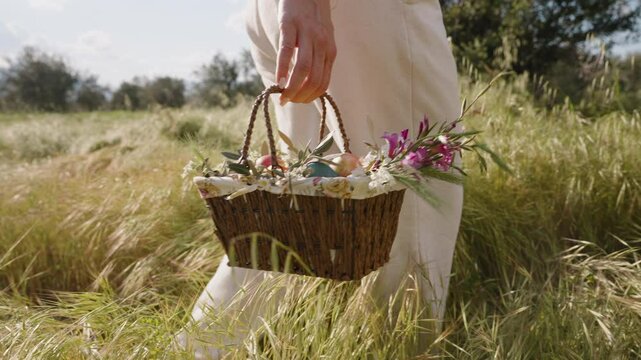 A Woman in White Pants Walks Through a Sunny Field, Holding a Woven Basket Filled with Colorful Flowers and Easter Eggs. Slow motion.
