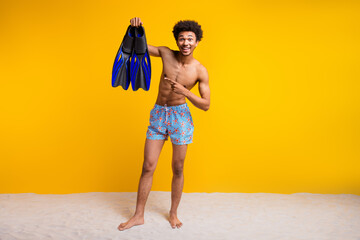 Happy young man in stylish swimwear holding snorkeling fins against a vibrant yellow background, embodying summer travel and leisure vibes © deagreez
