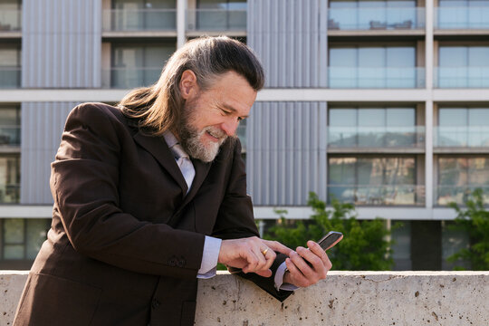 smiling middle age male with beard and gray hair in classy suit browsing on smartphone against modern city buildings, concept of business and mature man lifestyle, copyspace for text