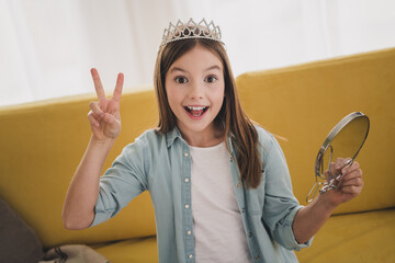 Delighted young girl wearing a tiara and denim shirt gesturing peace sign indoors © deagreez