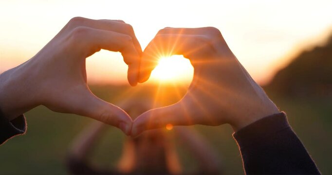 Man shows heart shape with hands to beautiful woman who does the same to him at sunset, slow motion. Close up of heart shape with the sunlight passing through the hands in sunset sky, love concept