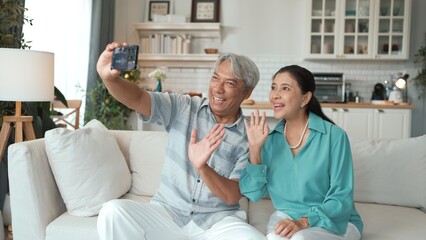 Obraz premium Happy grandfather and grandmother making video call with their son at home. Elder man holding smart phone and sitting at sofa while old woman greeting with cheerful vibe at modern house. Myrmidon.