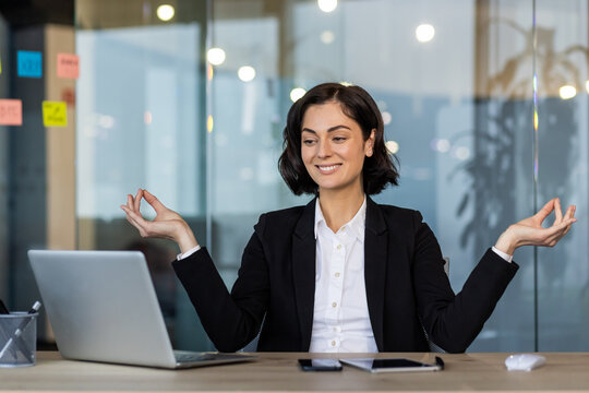 Businesswoman meditating in office at desk practicing mindfulness. She uses laptop for work tasks, creating calm focus and balance amidst busy workplace. Professional setting emphasizes career.